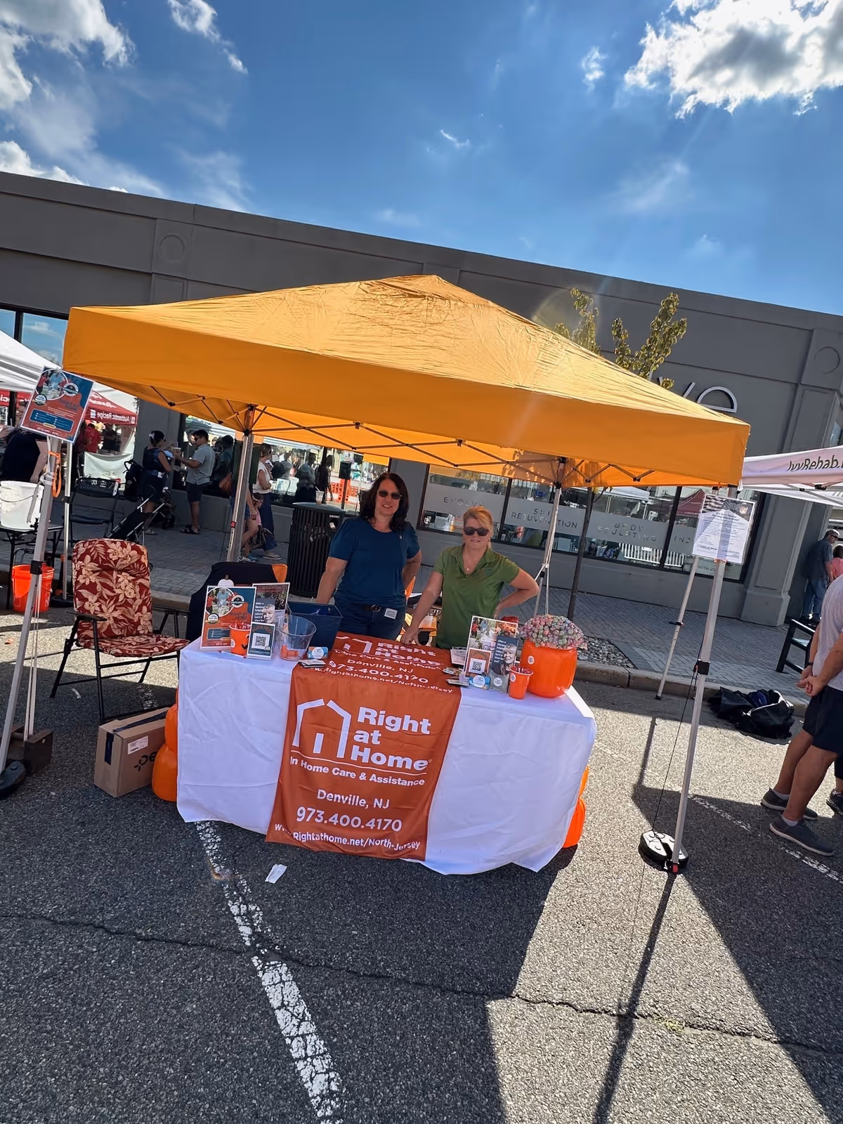 Two staff members stand behind an orange tent booth with Right at Home banner.