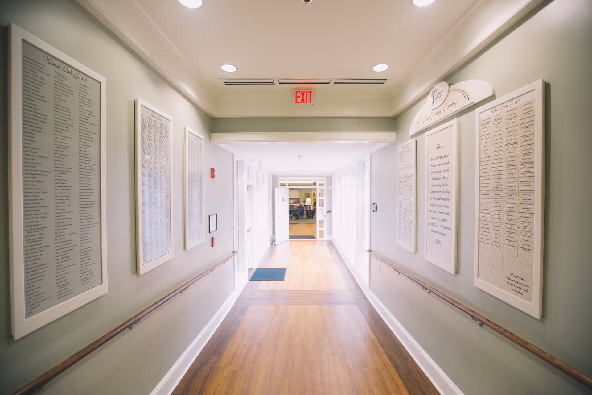 Well-lit hospice corridor with framed lists along pale green walls and an exit sign.