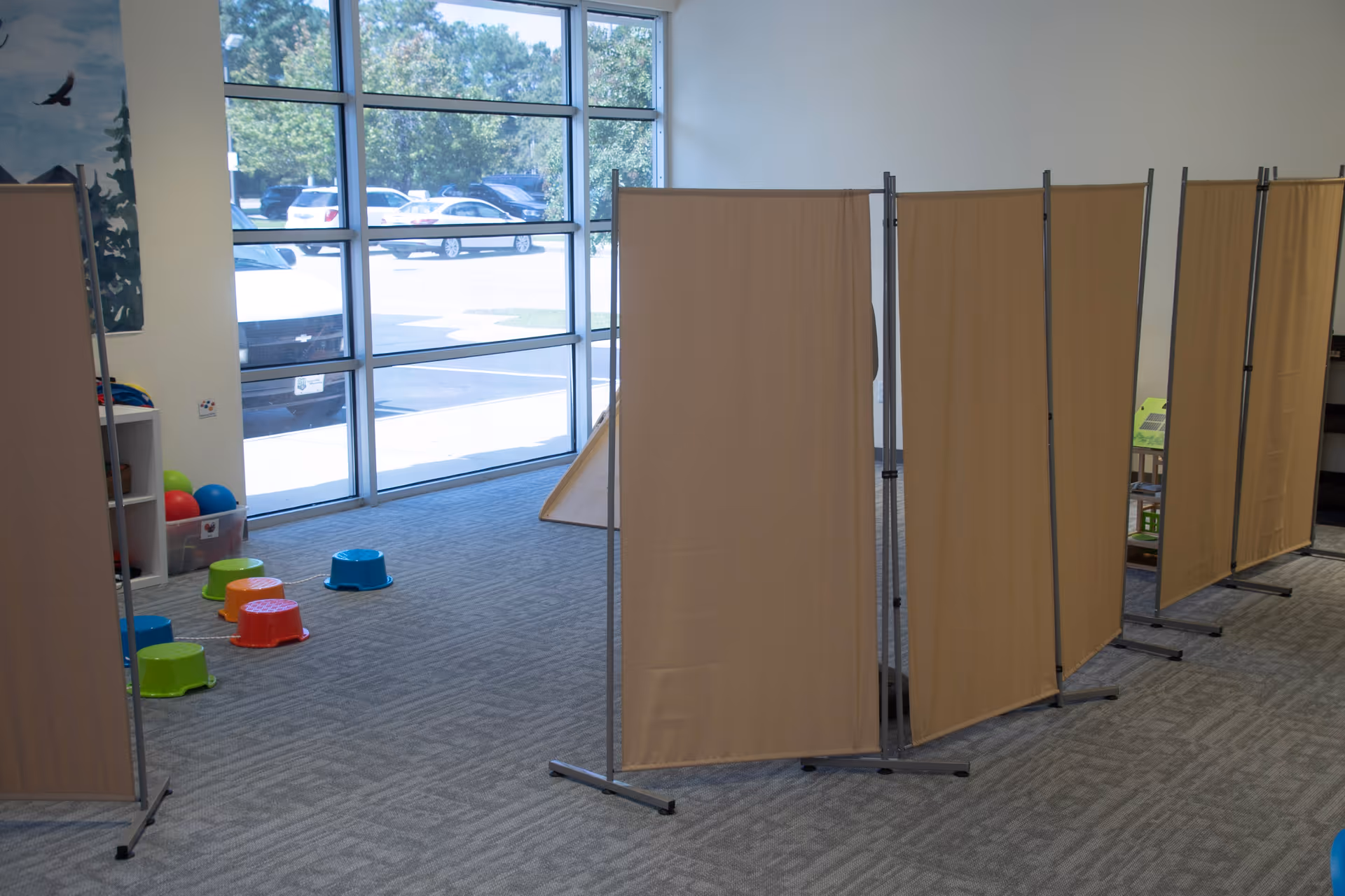 Interior space with beige folding room dividers, carpet, and colorful stools near large windows.