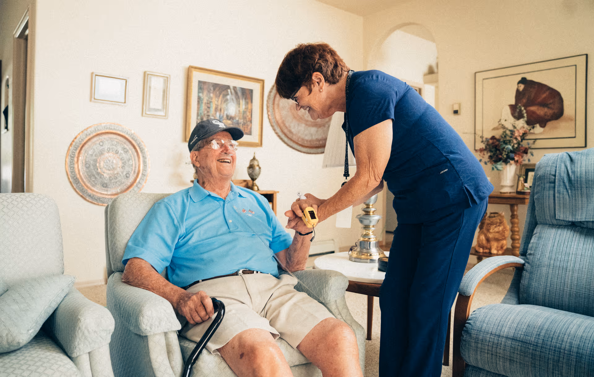 Caregiver in blue scrubs assists smiling elderly man seated in a living room.