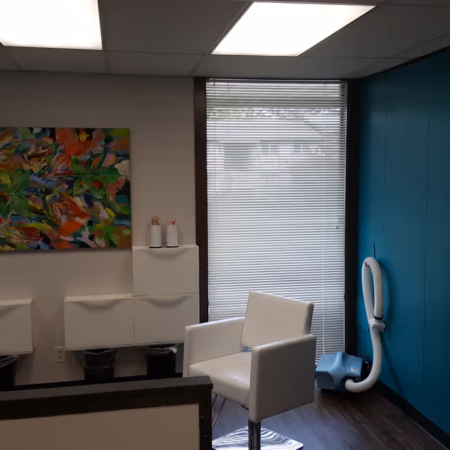 White modern chair beside teal wall, window blinds, colorful painting, and cleaning equipment.