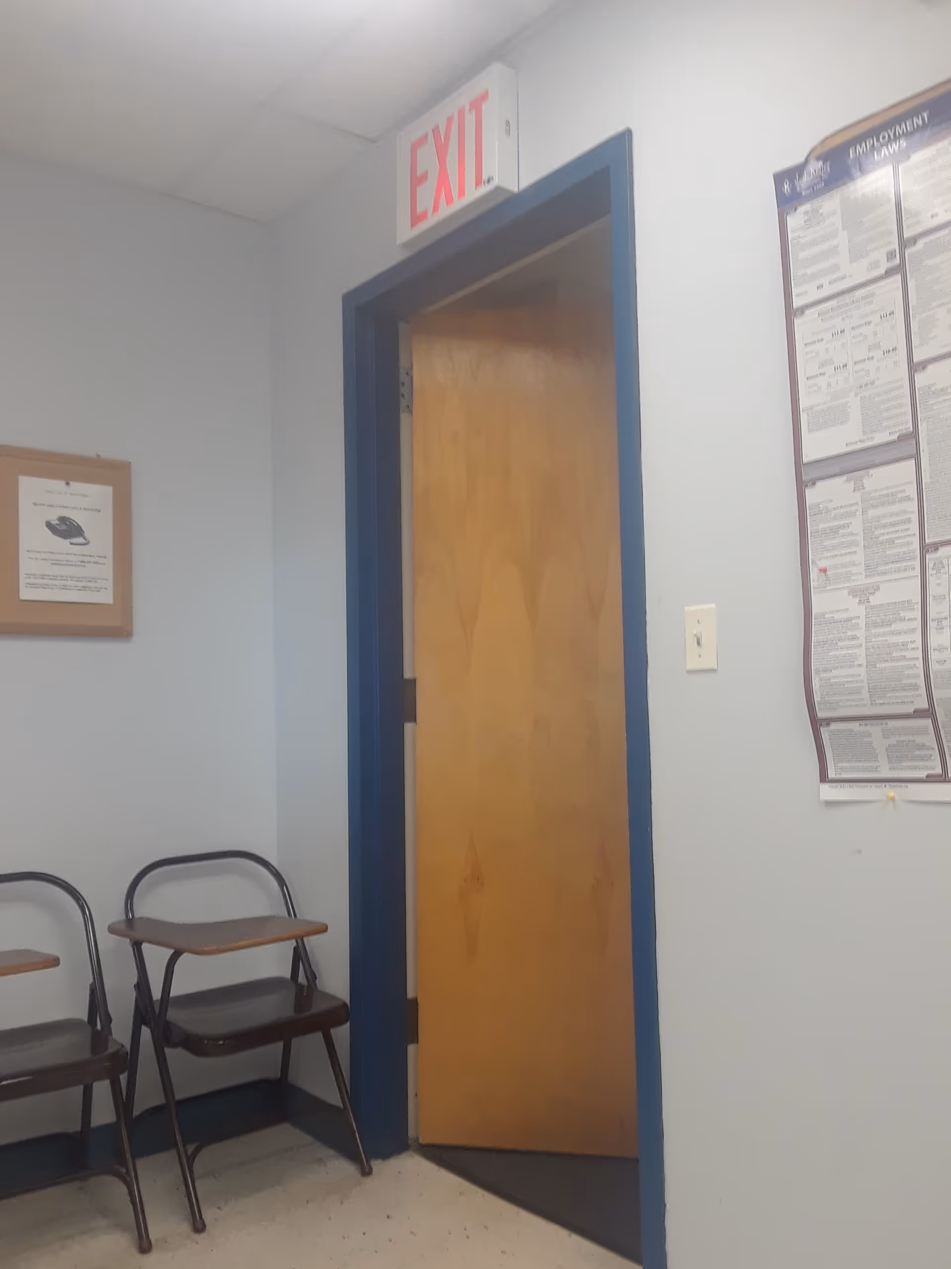 Interior hallway with open wooden door, exit sign, blue trim, and folding chairs.