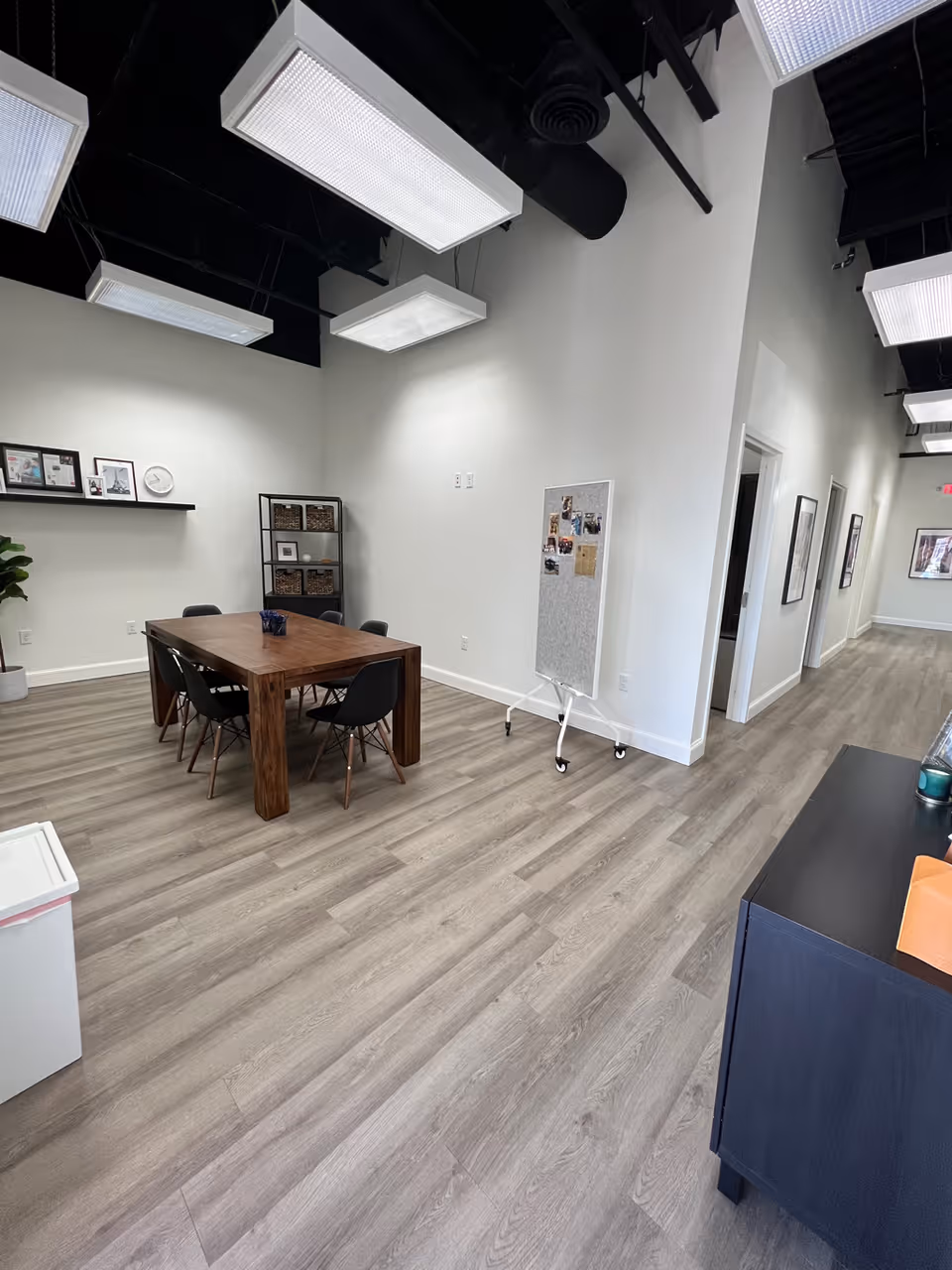 Interior communal room with a wooden table, chairs, shelving, and a mobile whiteboard.