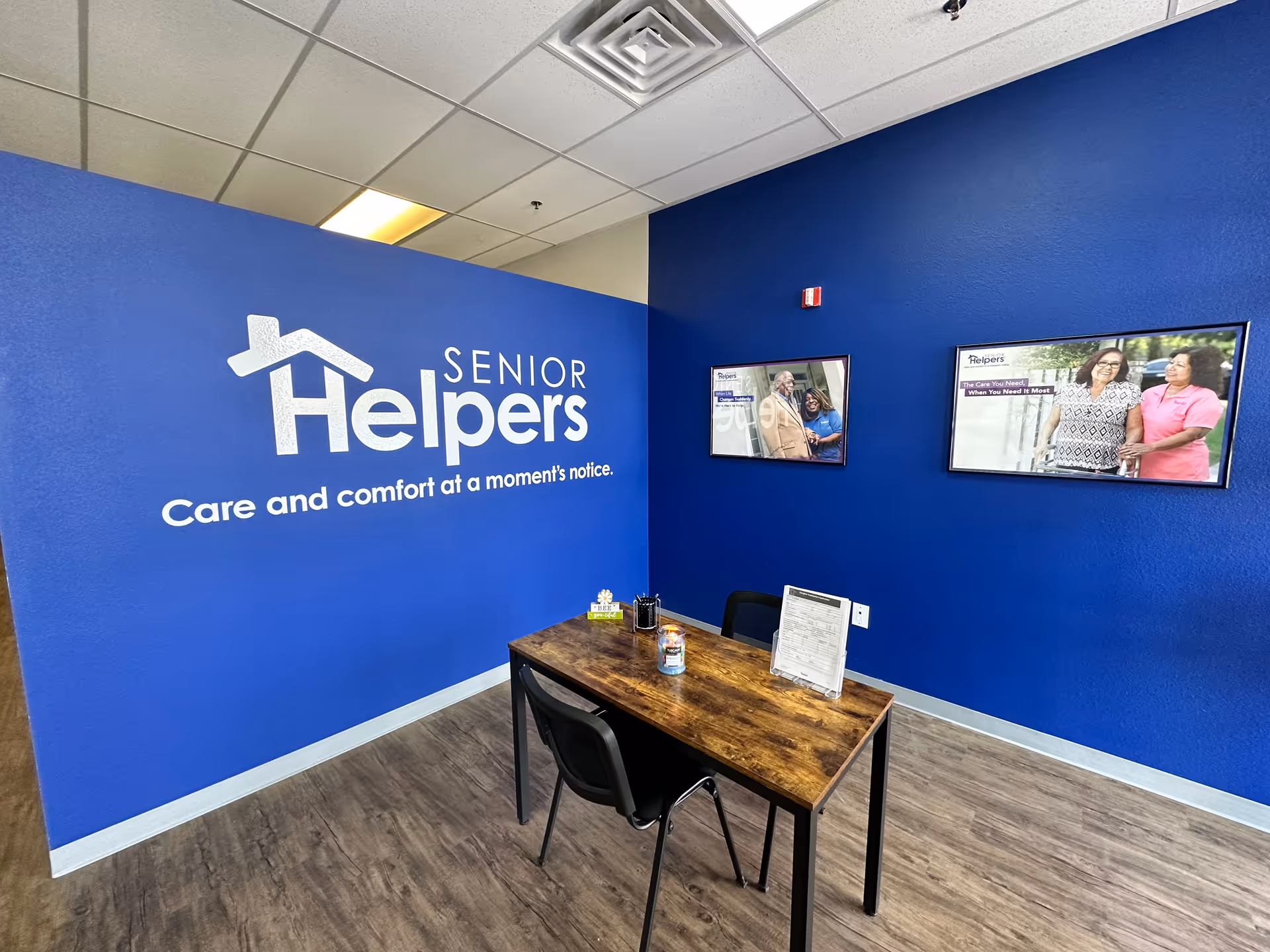Blue-walled office area with Senior Helpers logo, desk, chairs, and wall-mounted monitors.