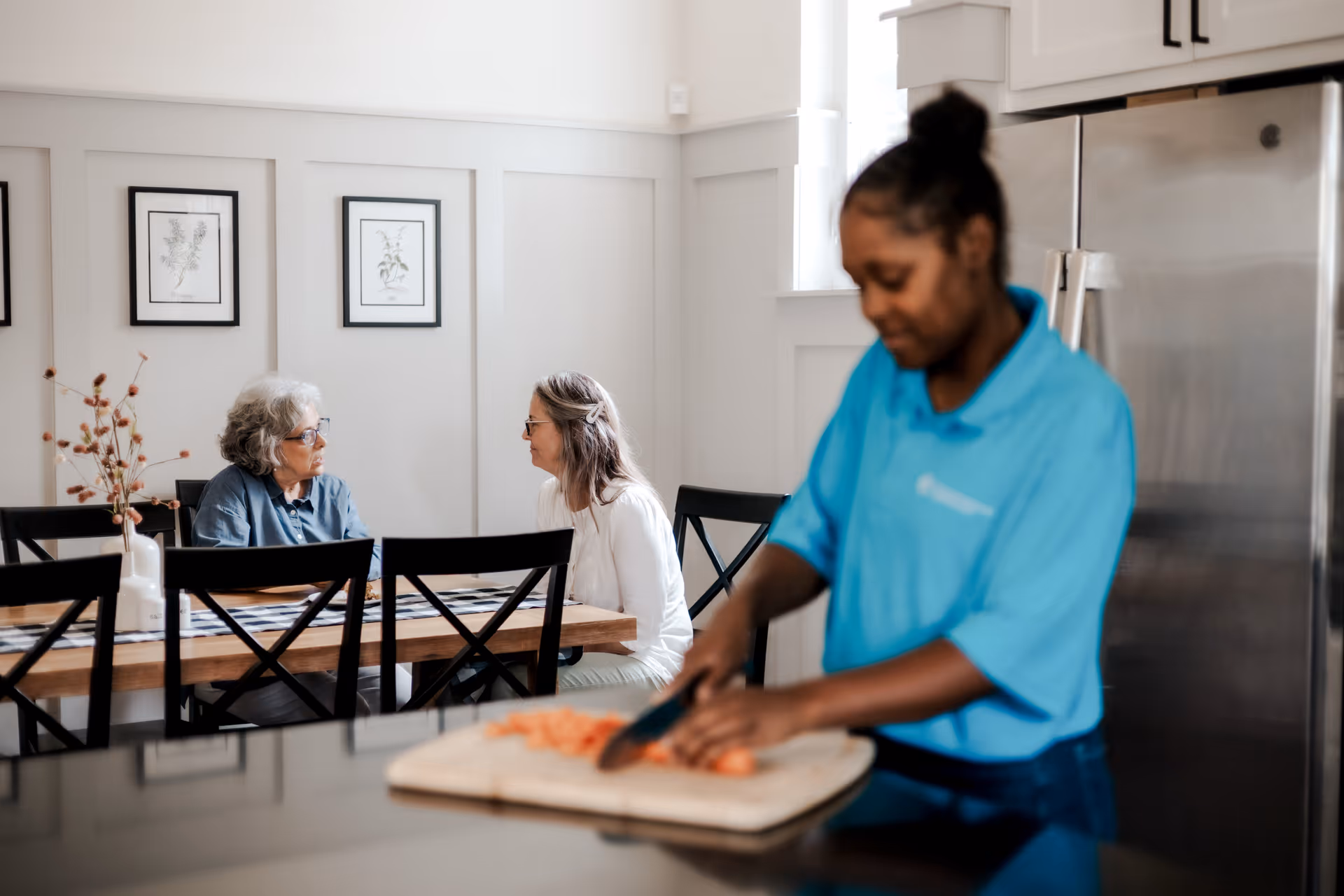 Two elderly residents converse at a dining table as a caregiver chops vegetables nearby.