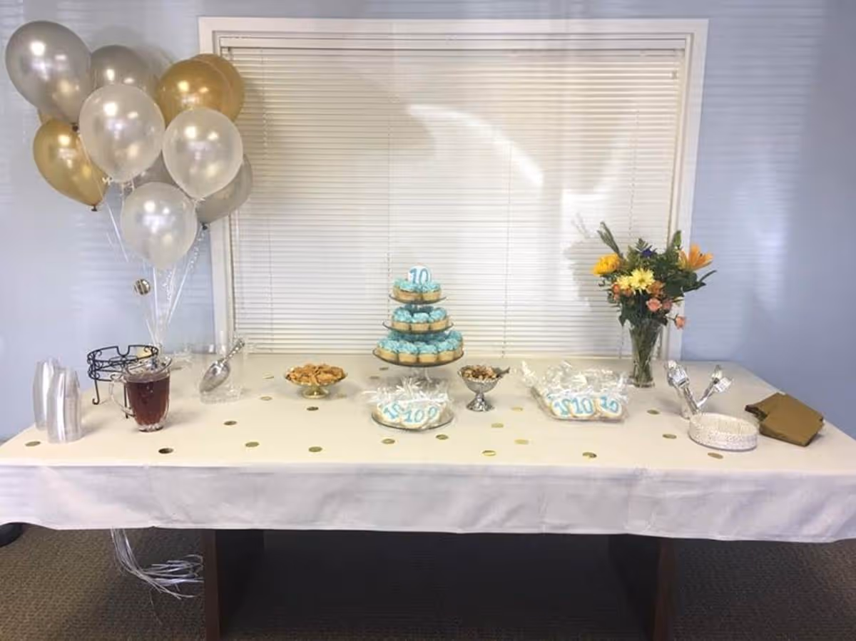 A decorated dessert table with balloons, cake, cookies, and a flower arrangement indoors.
