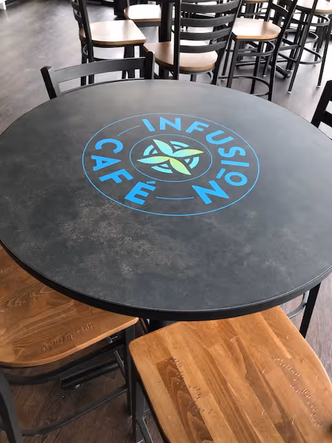 Round black cafe table with blue Infusion Café logo, indoors, surrounded by wooden chairs.