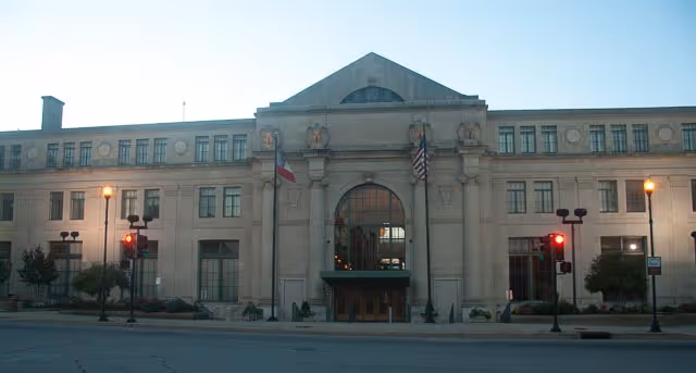 Grand stone building with an arched entrance, flags, and street lamps along a city street.