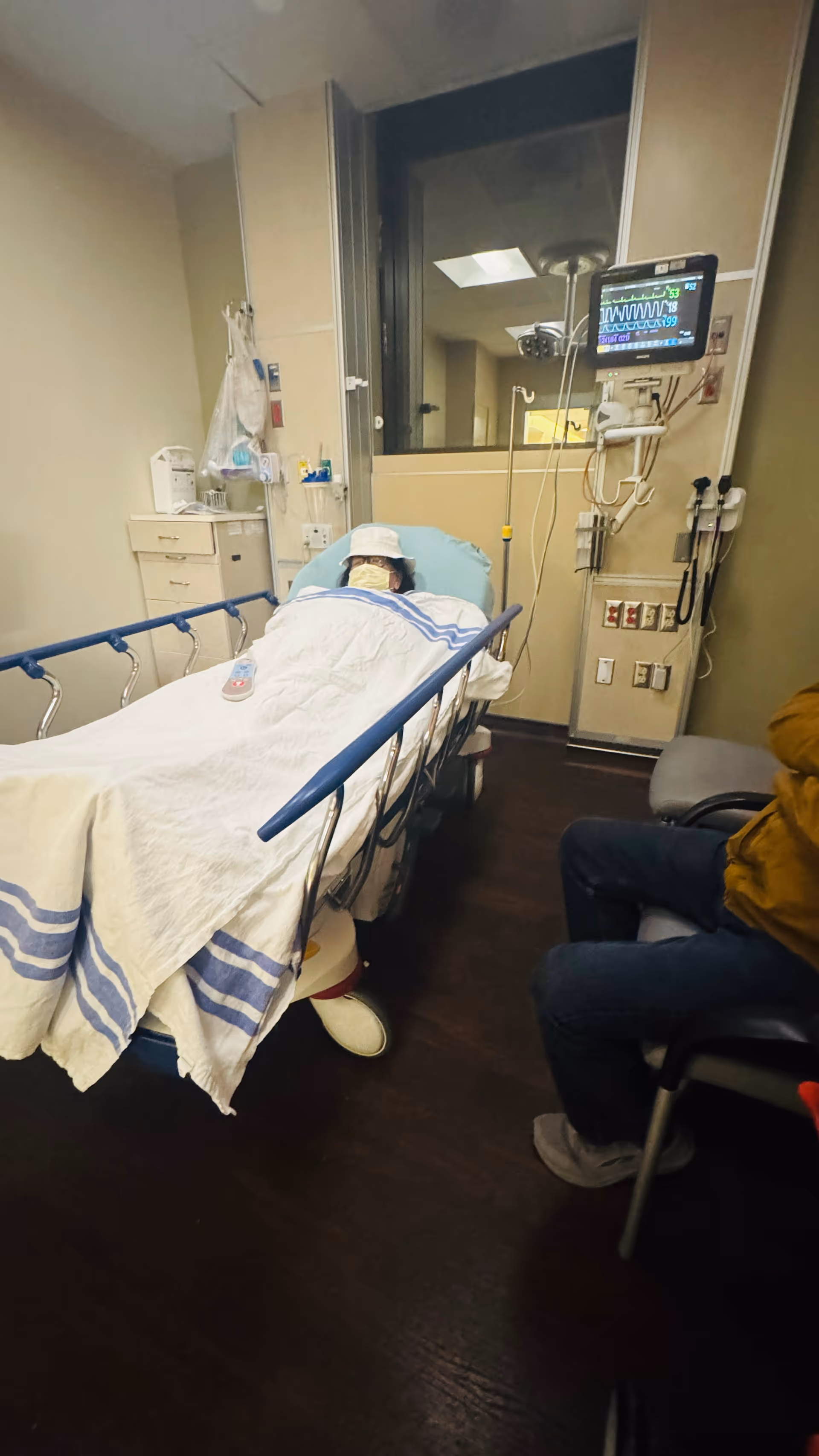 Hospital emergency room bed with patient, monitoring equipment, and a nearby chair.