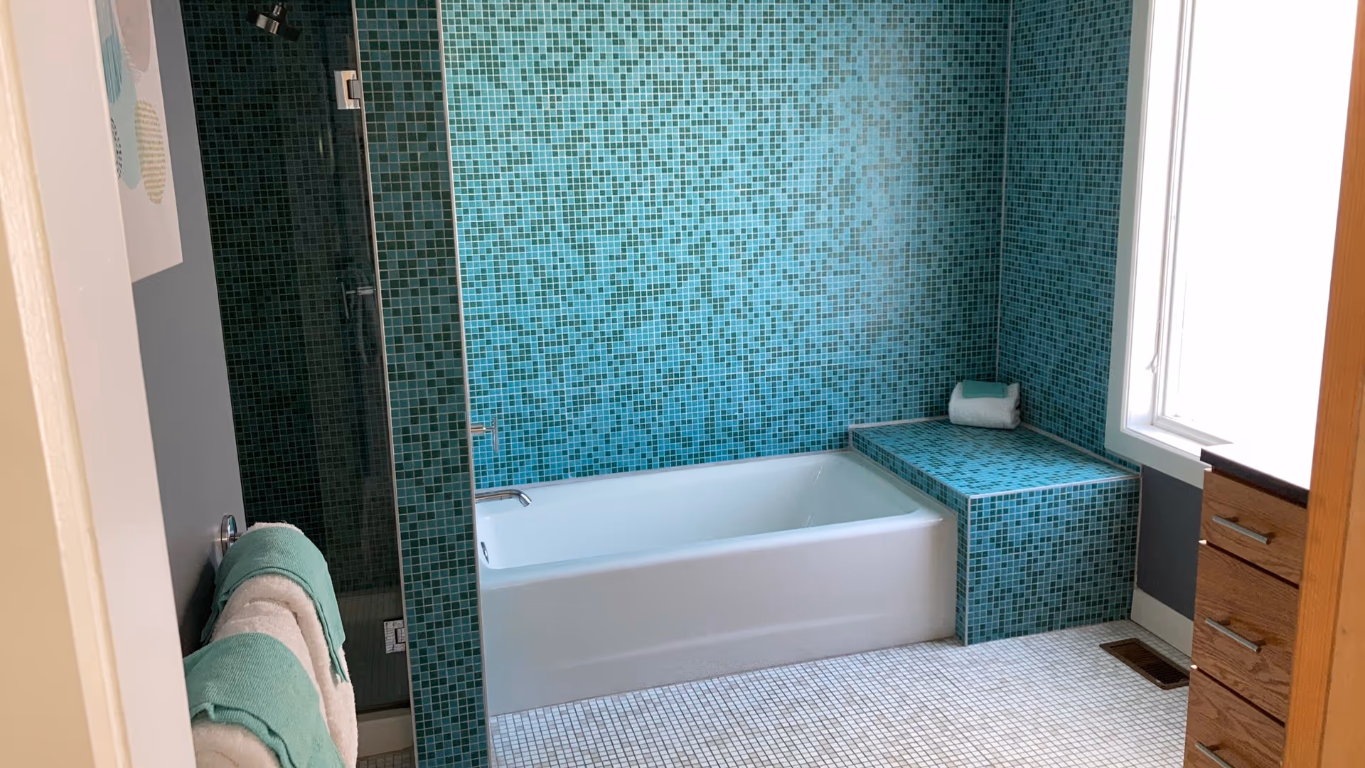 Modern turquoise-tiled bathroom featuring a bathtub, shower, and window.