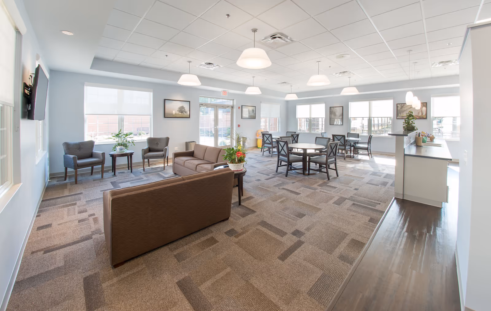 Bright senior living lounge with brown sofas, chairs, tables, and large windows.
