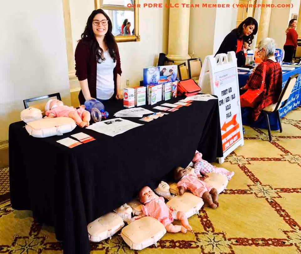 Smiling woman stands at a CPR training booth with mannequins and brochures.