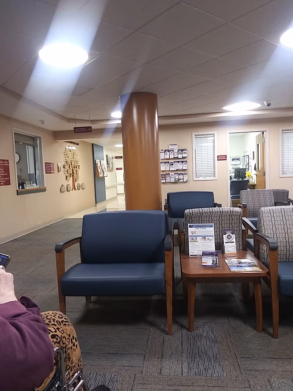 A hospital lobby with blue chairs, wooden tables, brochures, and a central column.