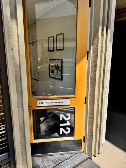 Orange-framed glass door labeled 212 leads to Newport Home Care hallway with framed pictures.