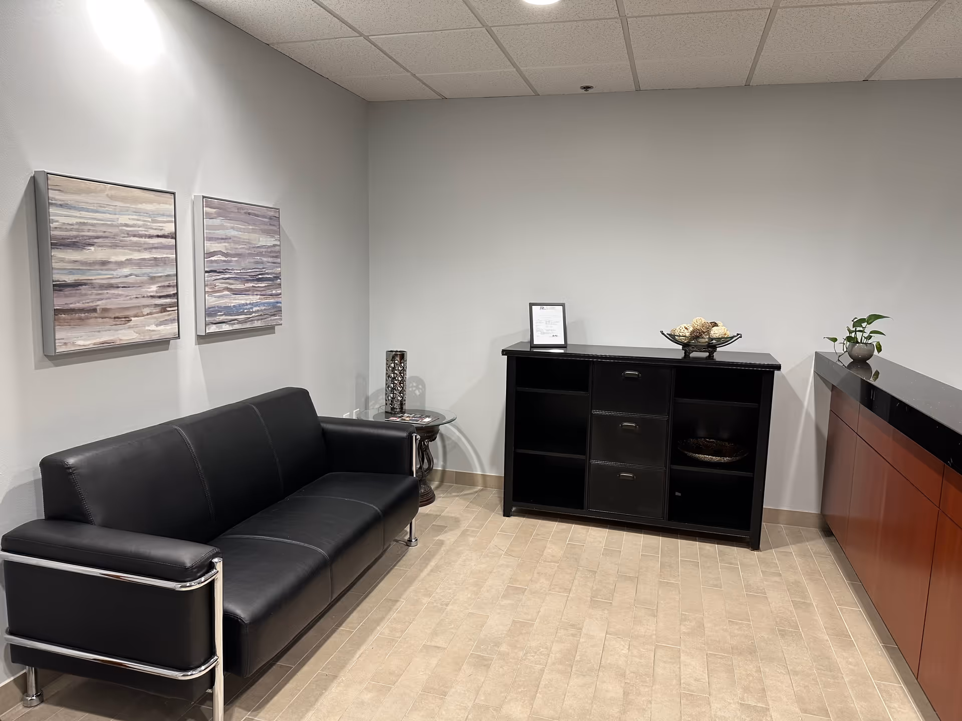 Modern waiting area with black leather sofa, artwork, and a dark sideboard against a gray wall.