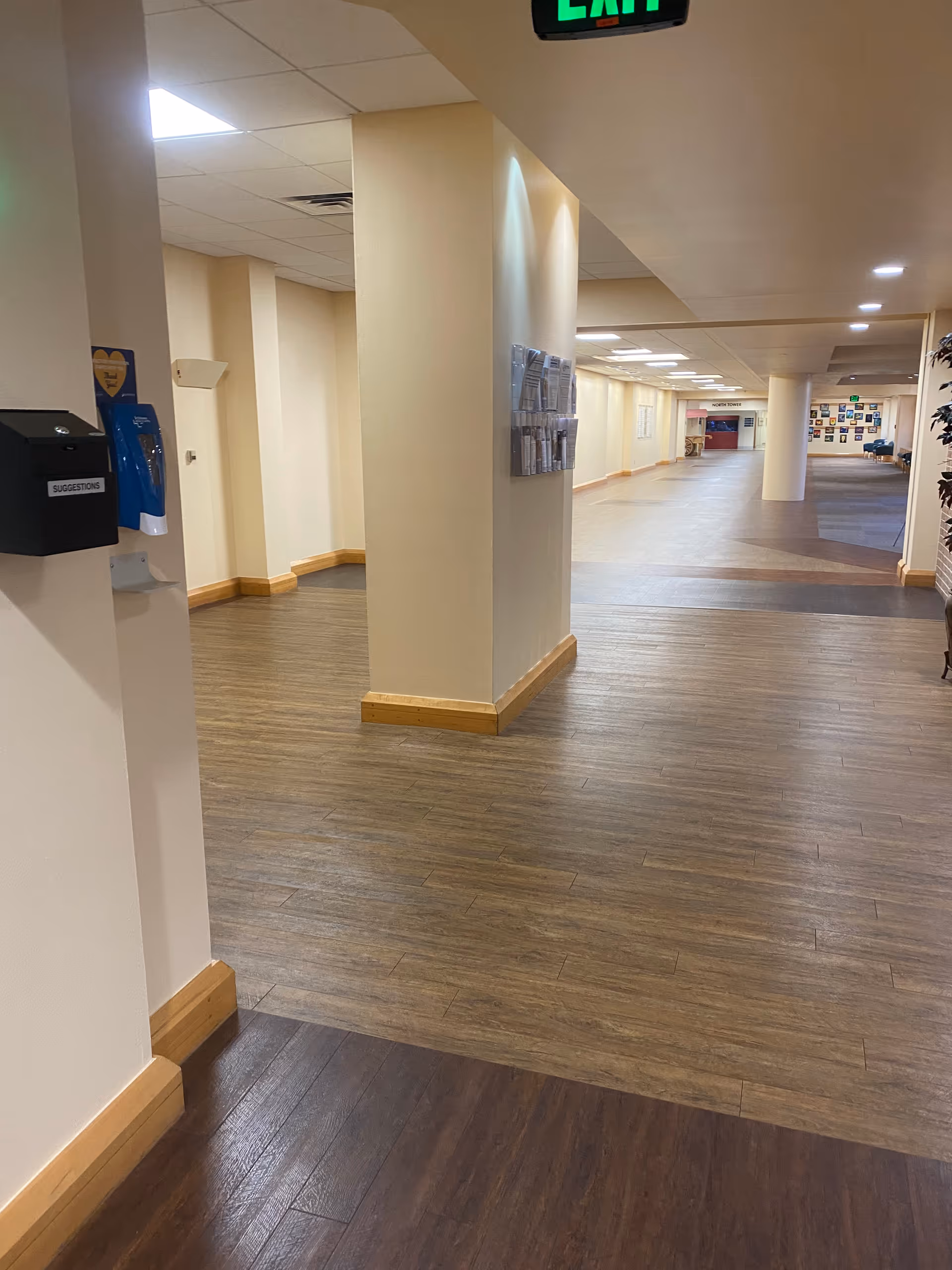 Long, well-lit hospital hallway with columns, bulletin boards, and wood-style flooring.