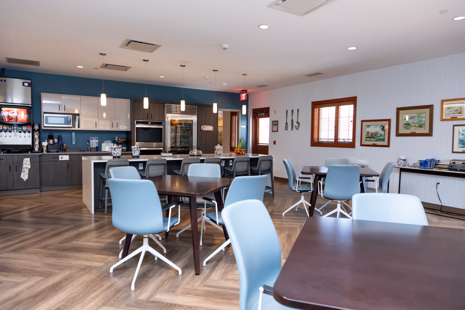 Modern dining area with blue chairs, wooden tables, and a kitchen/service counter in the background.
