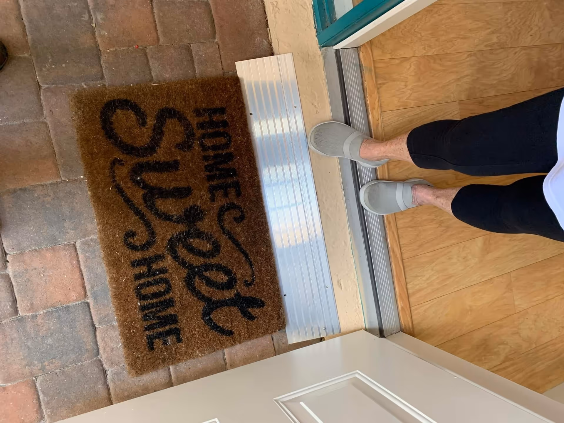 Person stands at interior doorway, gray slippers beside a 'Home Care' welcome mat.