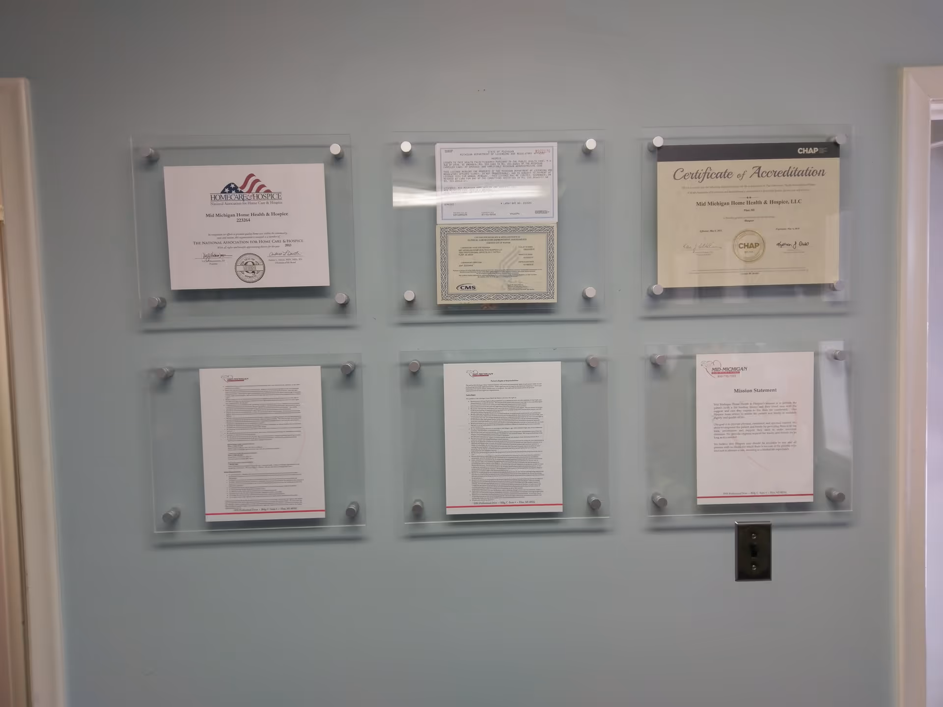 Six glass-framed certificates arranged on a pale green wall in a hallway.