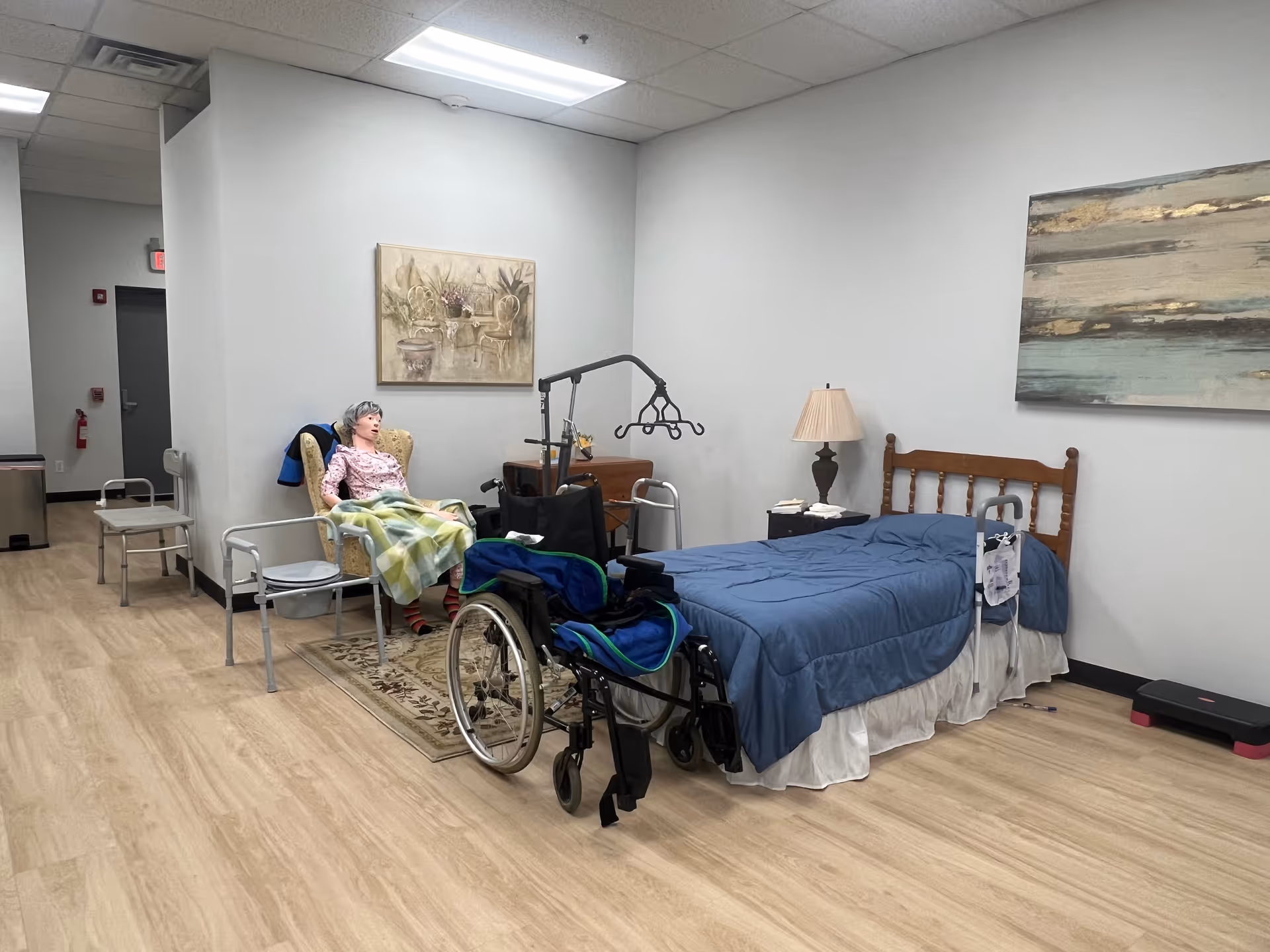 Elderly resident seated in a chair beside a hospital-style bed in a care room.
