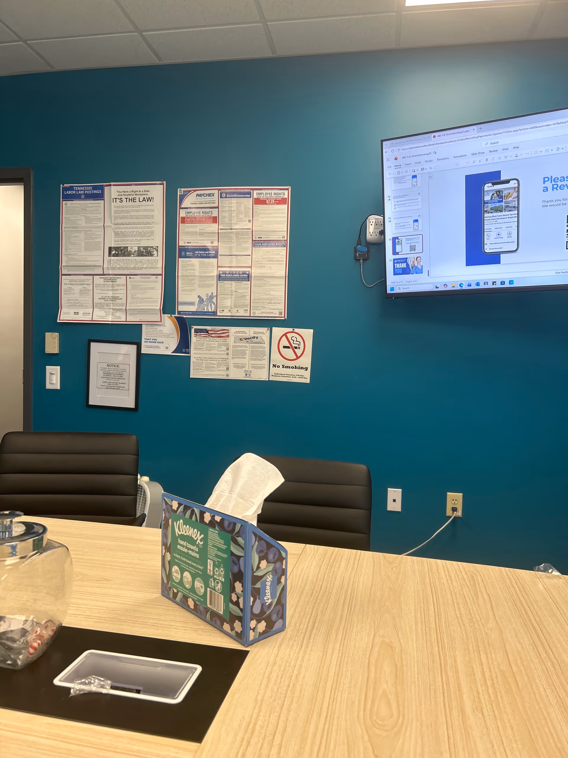 Teal-walled conference area with bulletin boards, mounted screen, and a wooden table.