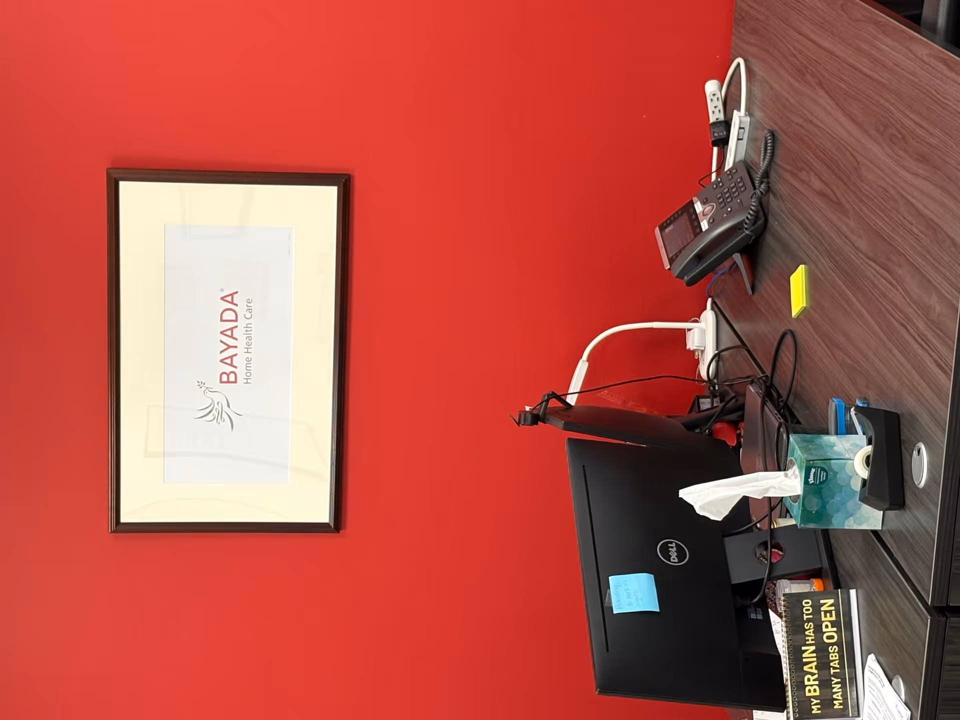 Office desk against red wall with Dell computer, phone, tissues, and framed BAYADA certificate.