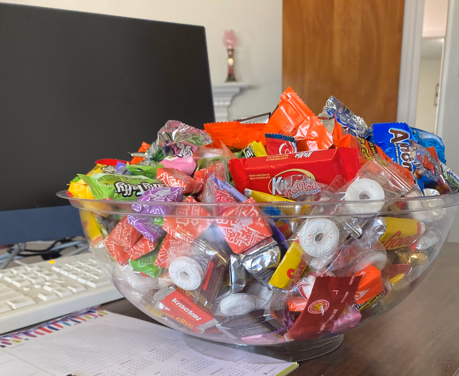 Glass bowl overflowing with assorted wrapped candies on a desk in an office.