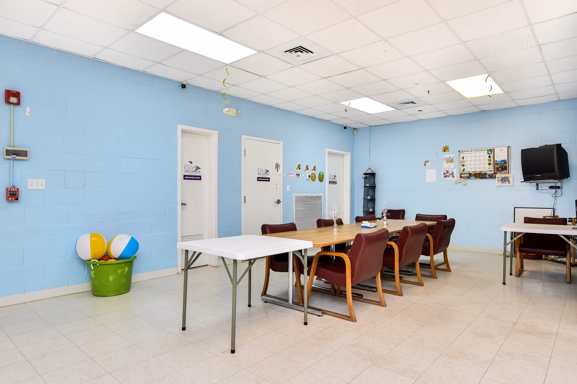 Blue-walled indoor common area with a long table, chairs, TV, and bulletin board.