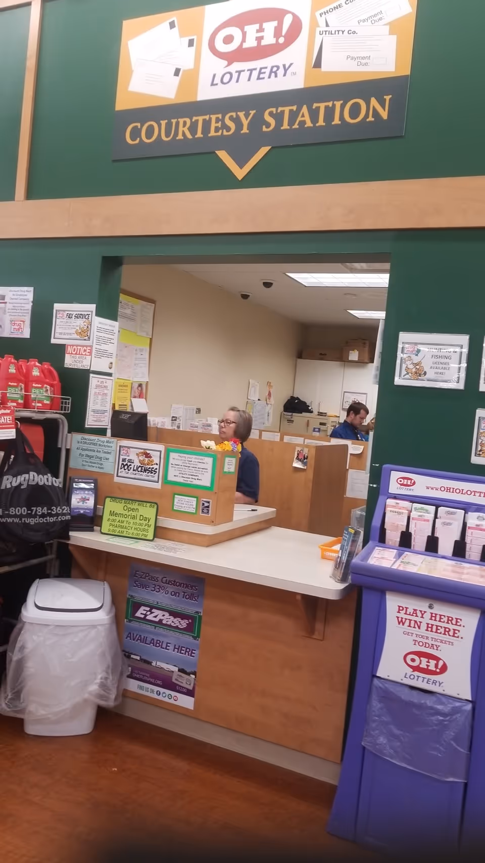 Inside Discount Drug Mart, a courtesy station counter with staff, signs, and a purple lottery stand.