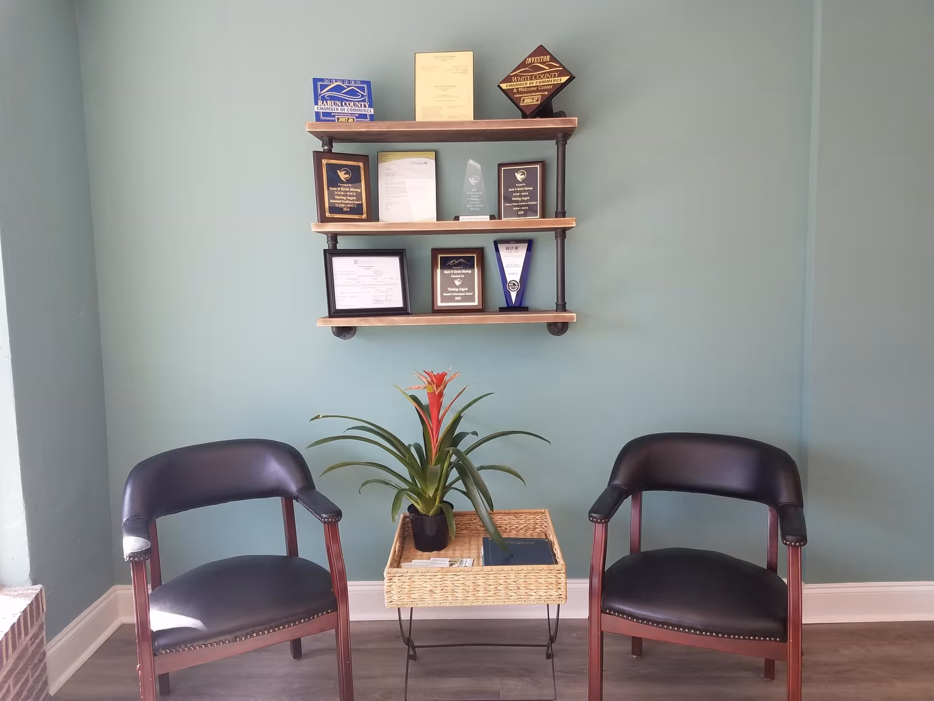 Two dark chairs flank a small wicker table with a plant in a teal-walled waiting area.