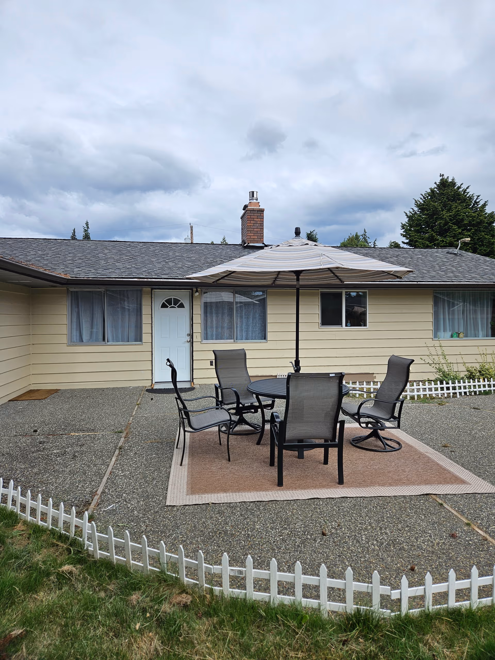 Outdoor patio with table, chairs, umbrella, and white picket fence in front of a single-story home