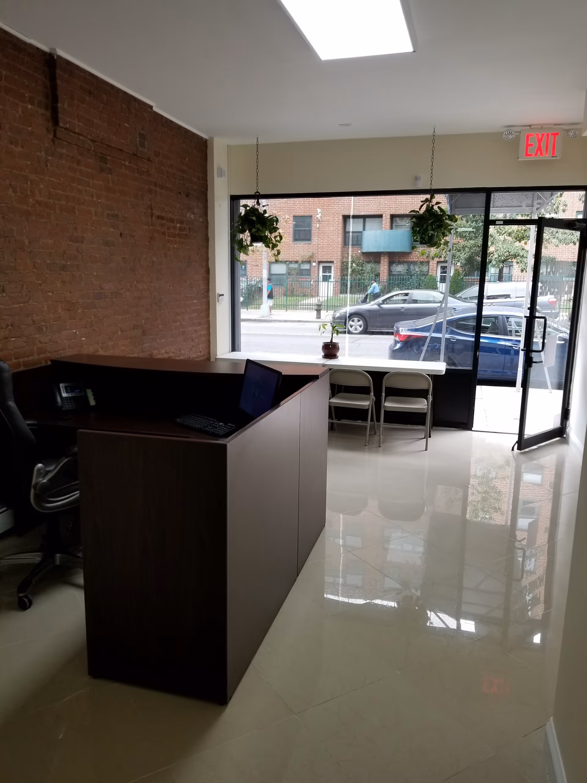 Reception area with dark wood desk, brick wall, large front windows, and plants.