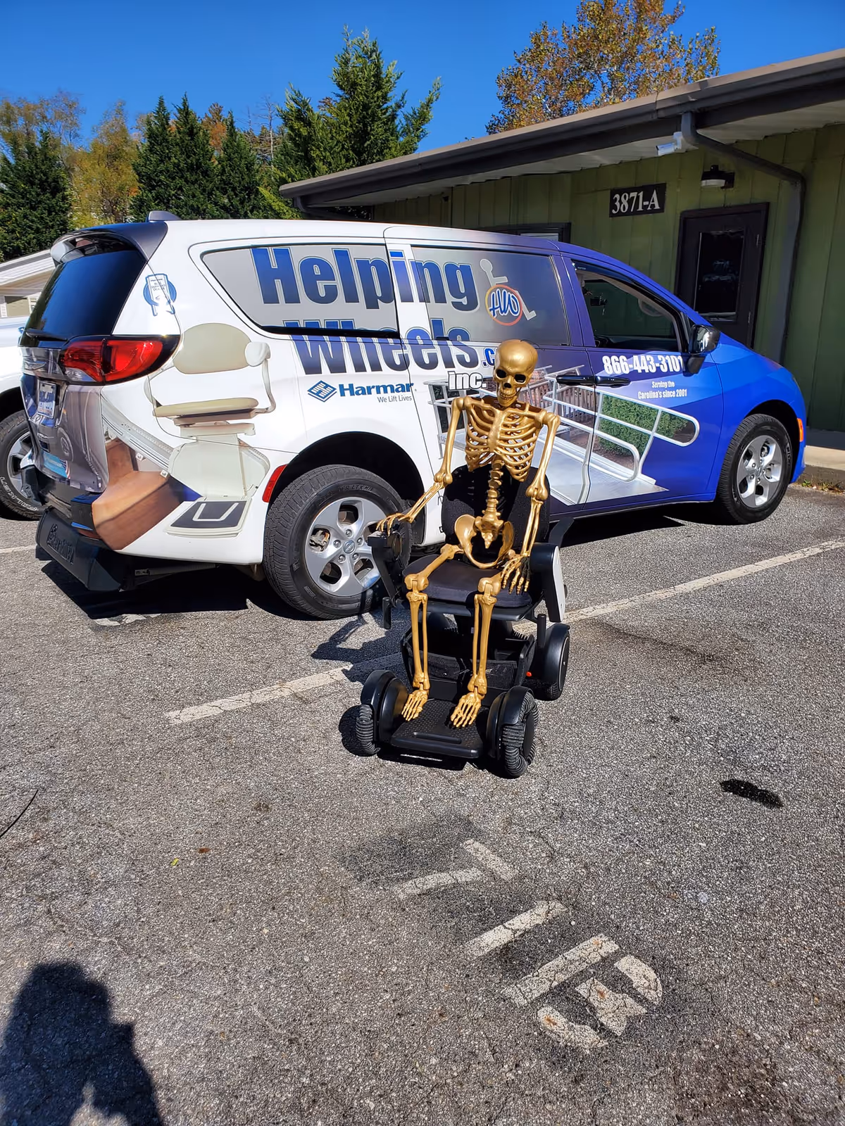 A Helping Wheels Inc van parked outside a building with a skeleton on a mobility scooter.