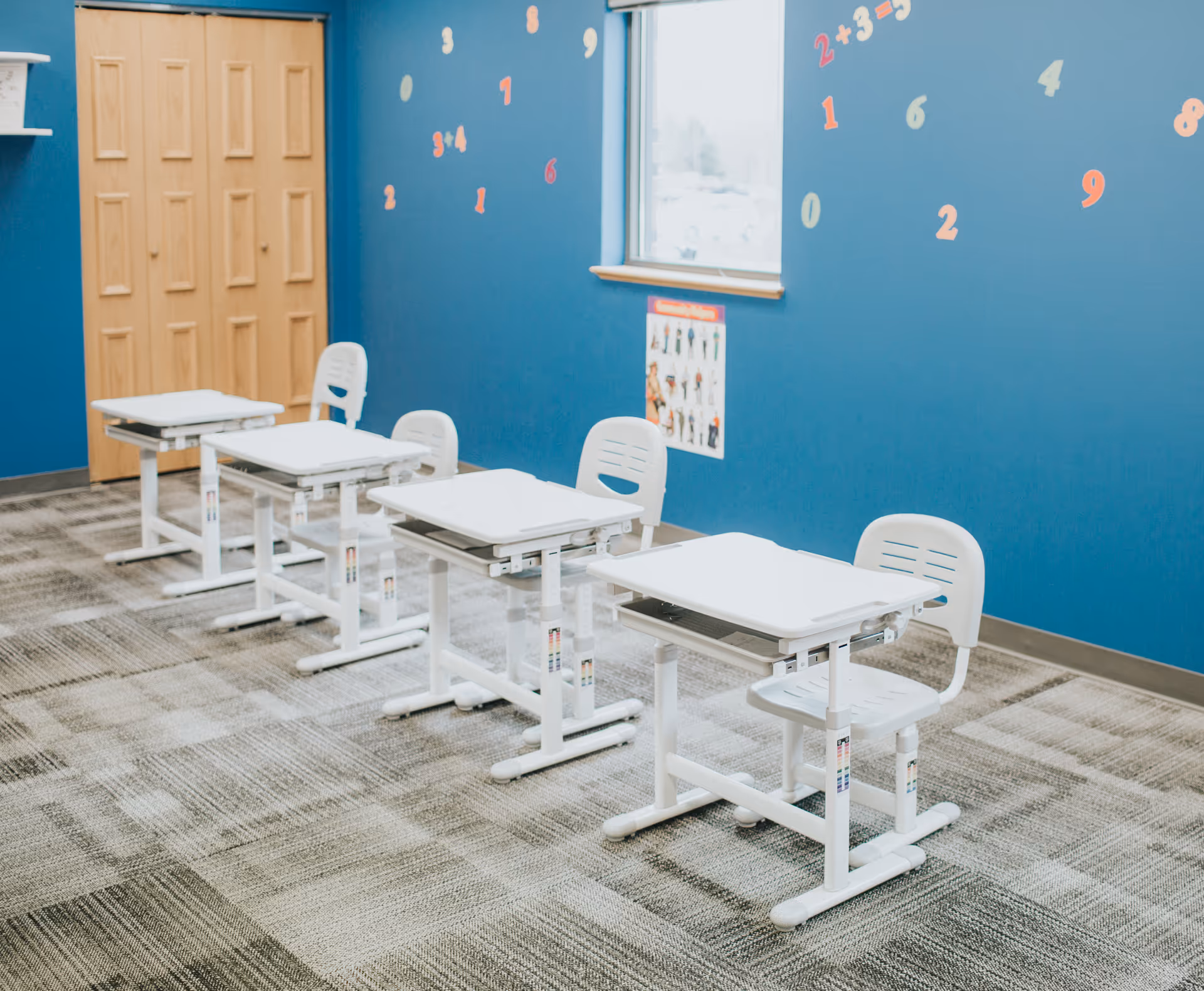 Bright blue classroom-like room with white desks and chairs arranged in a row.