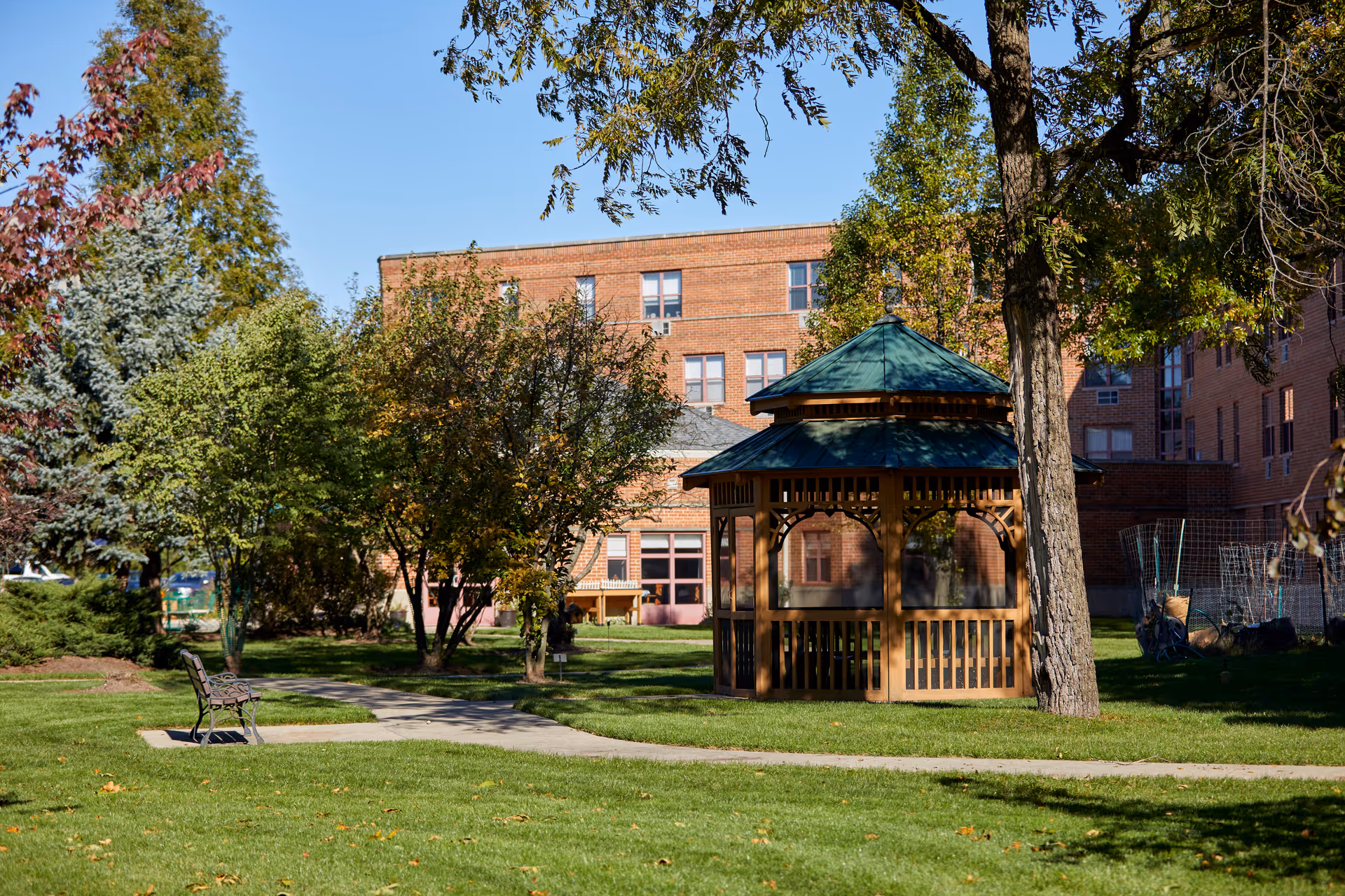 Brick senior living building with a gazebo in a grassy courtyard shaded by trees.