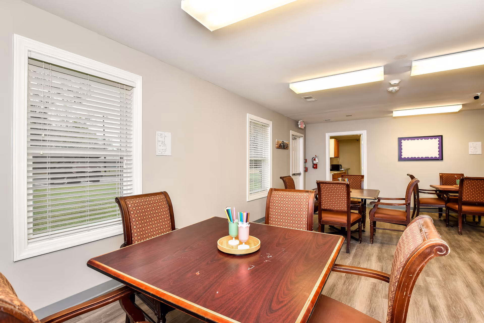 Senior living dining room with brown tables, upholstered chairs, and bright overhead lights.