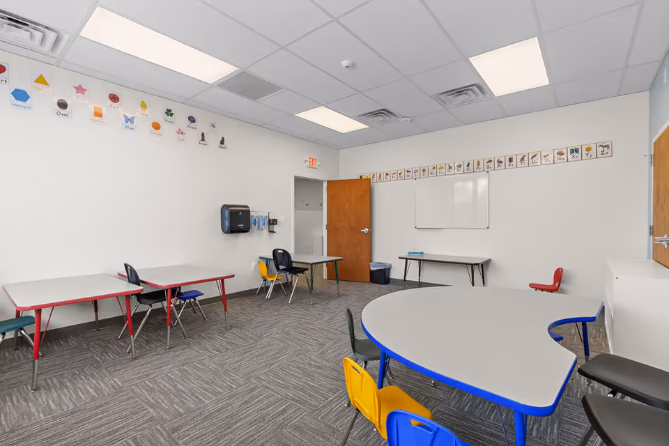 Interior classroom-style room with rounded table, small chairs, whiteboard, and colorful wall posters.