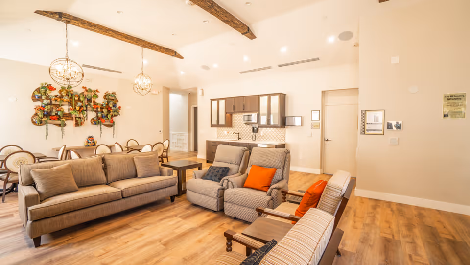 Bright communal living area with sofas, chairs, and a kitchenette visible in the background.