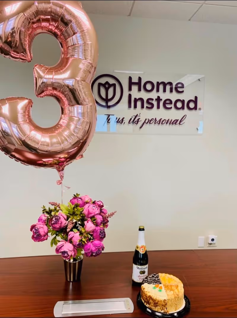 Indoor celebration scene at Home Instead with a balloon, flowers, champagne and cake.