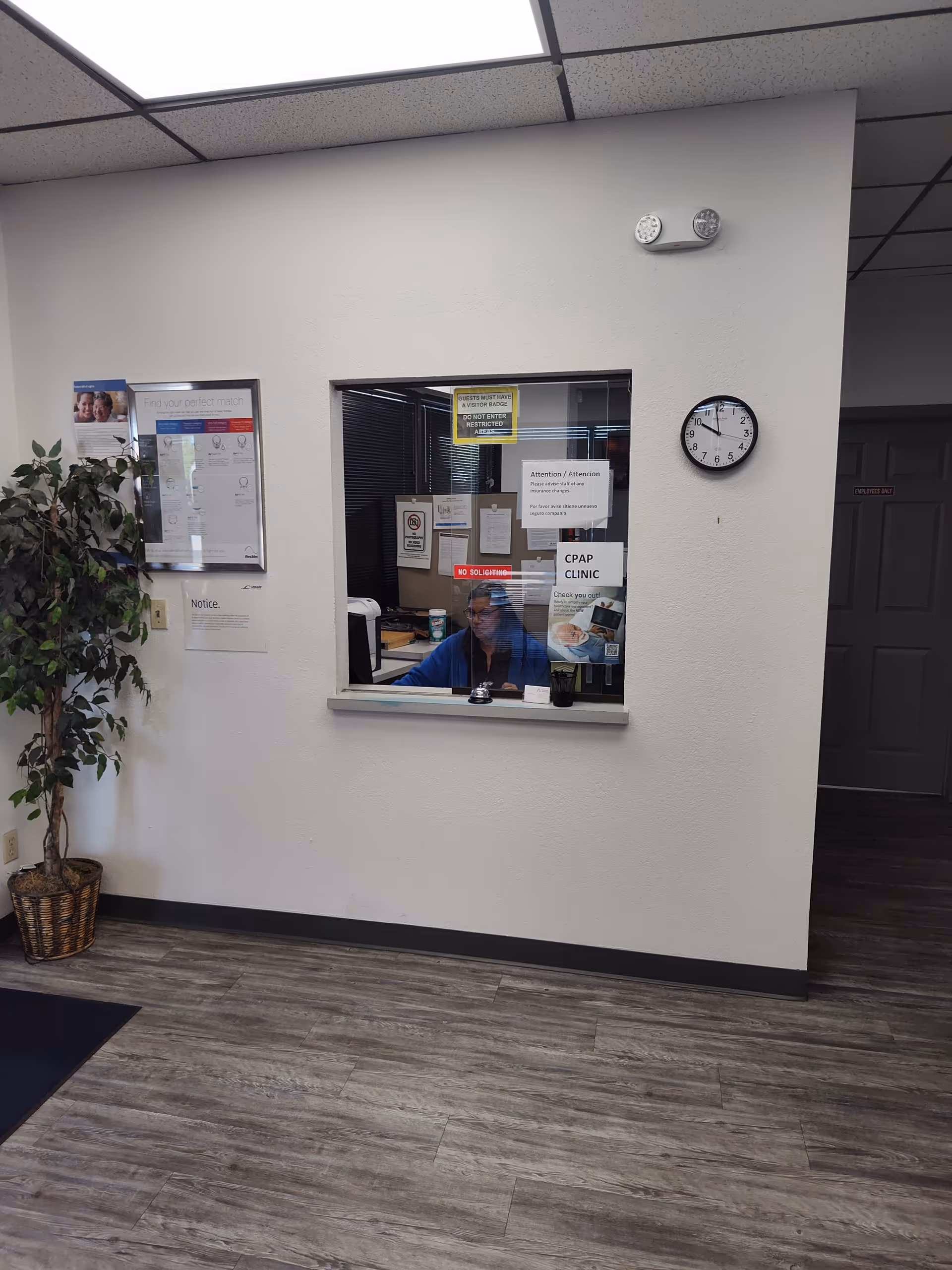 Reception area with front desk window, wall clock, potted plant, and staff member.