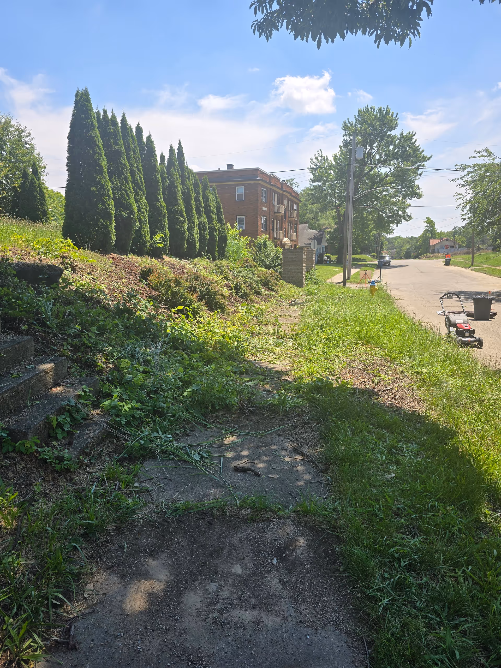Brick building along a grassy street with tall evergreen shrubs and a lawn mower.