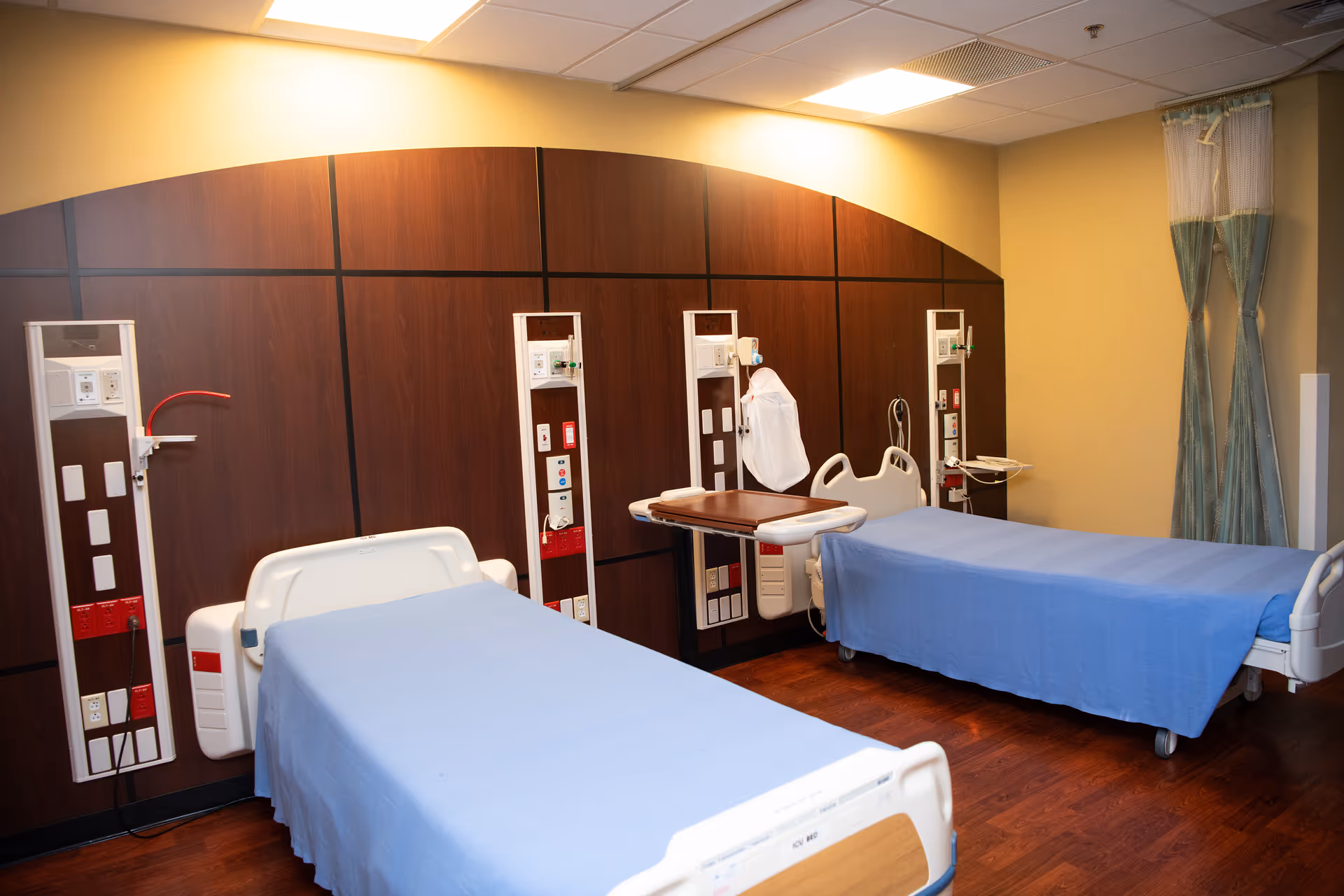 Two hospital beds with blue sheets in a wood-paneled patient care room.