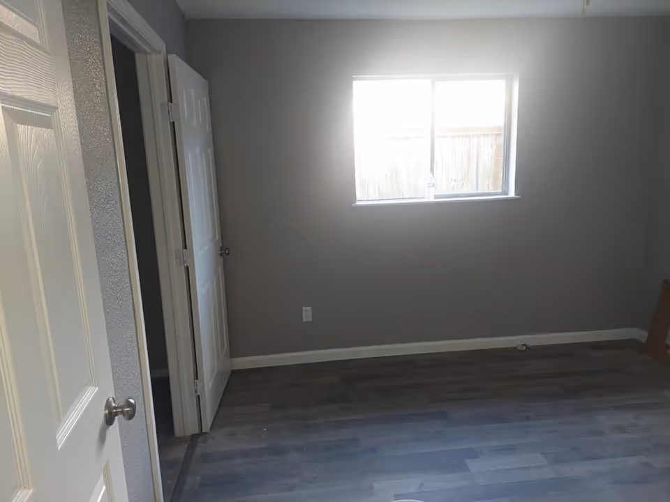 Empty gray-walled room with a window, doors, and dark wood flooring.