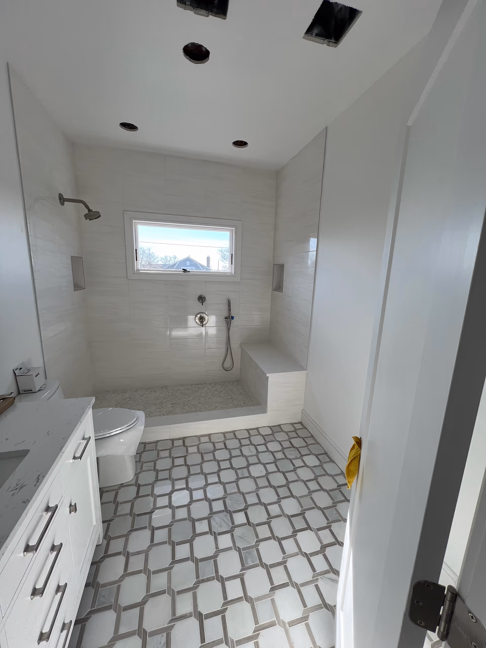 Bright white bathroom with a shower, built in bench, small window, and patterned floor tiles.