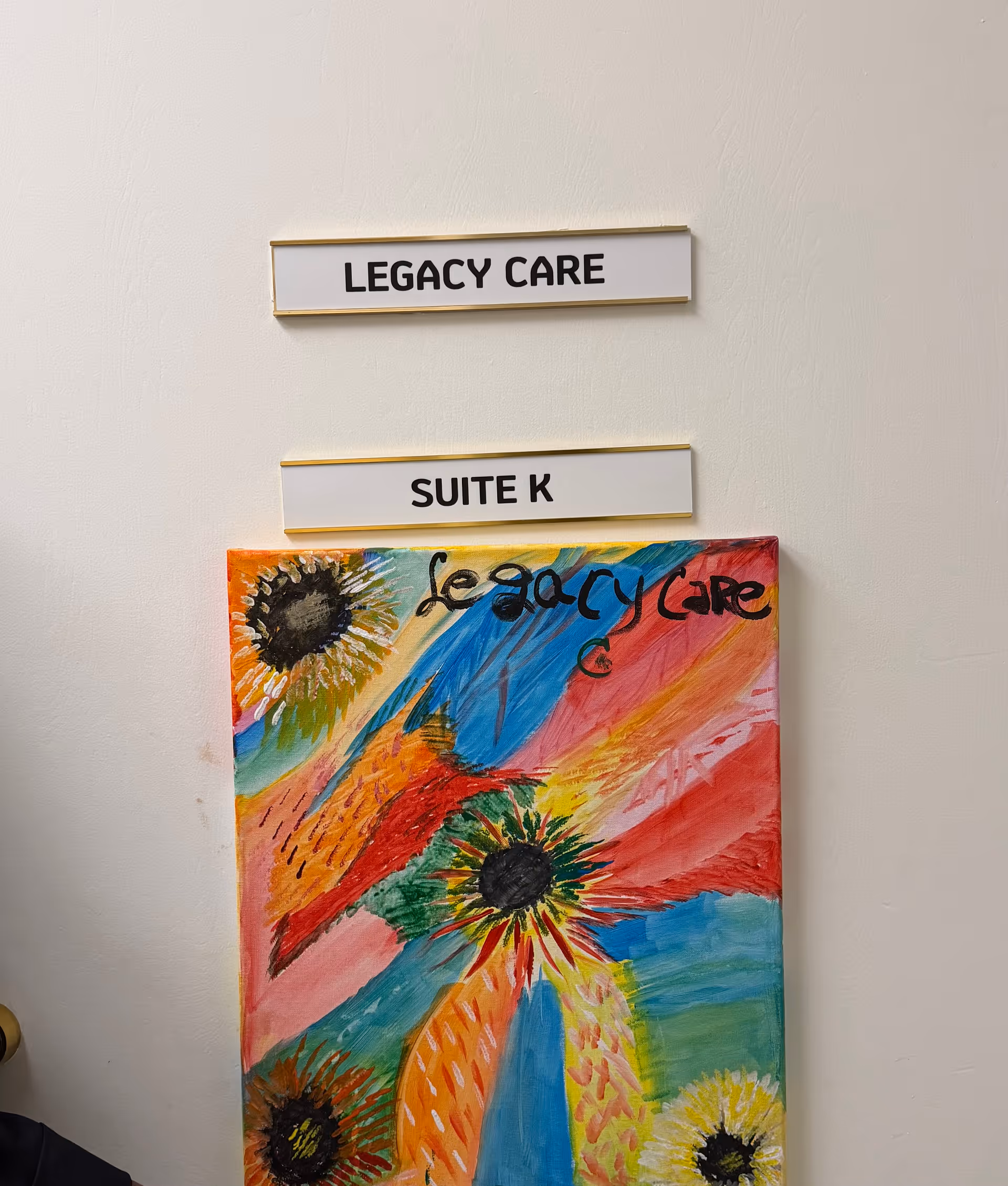 Interior wall with Legacy Care sign, Suite K plaque, and a colorful abstract painting.