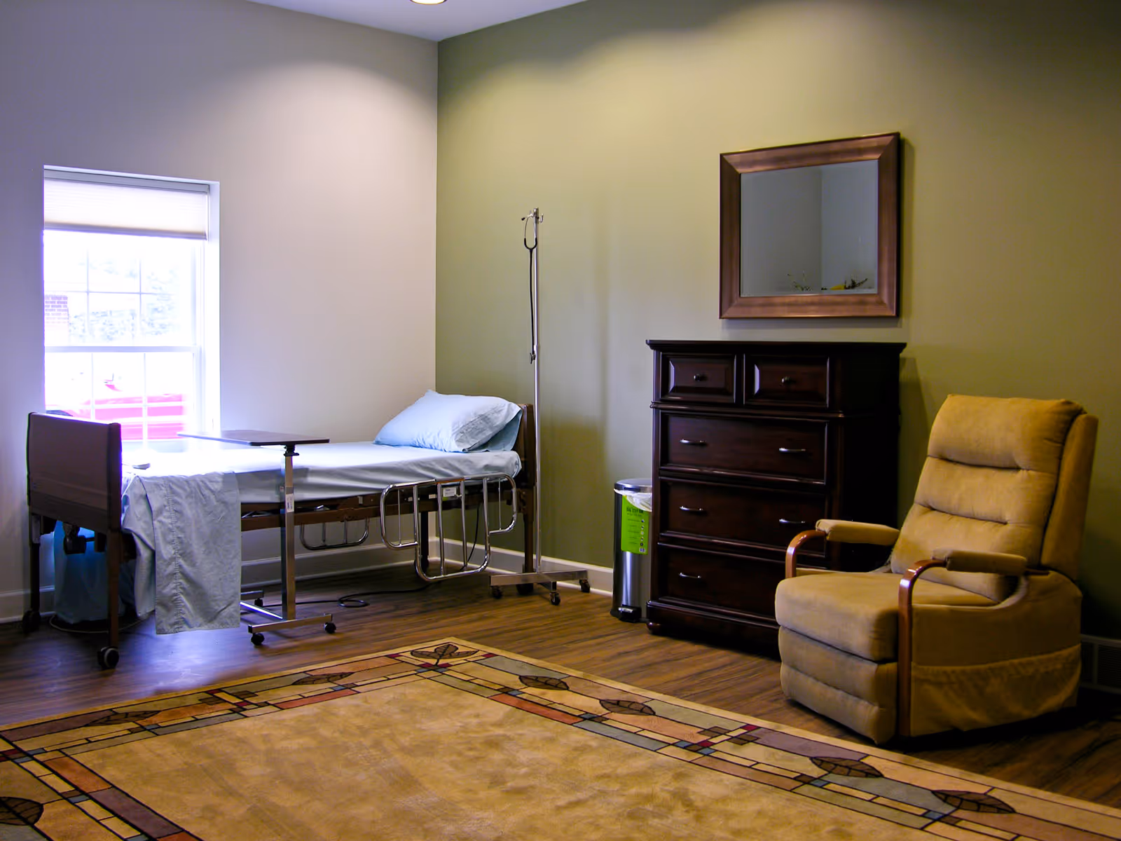 A bedroom in a senior living facility featuring a hospital-style bed, IV pole, dresser, and recliner.