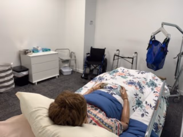 Resident rests on a floral bed in a small, medical-equipped bedroom with mobility aids.