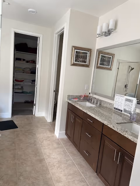 Bathroom vanity area with granite countertop, double sinks, mirrors, light fixtures, and open linen closet.