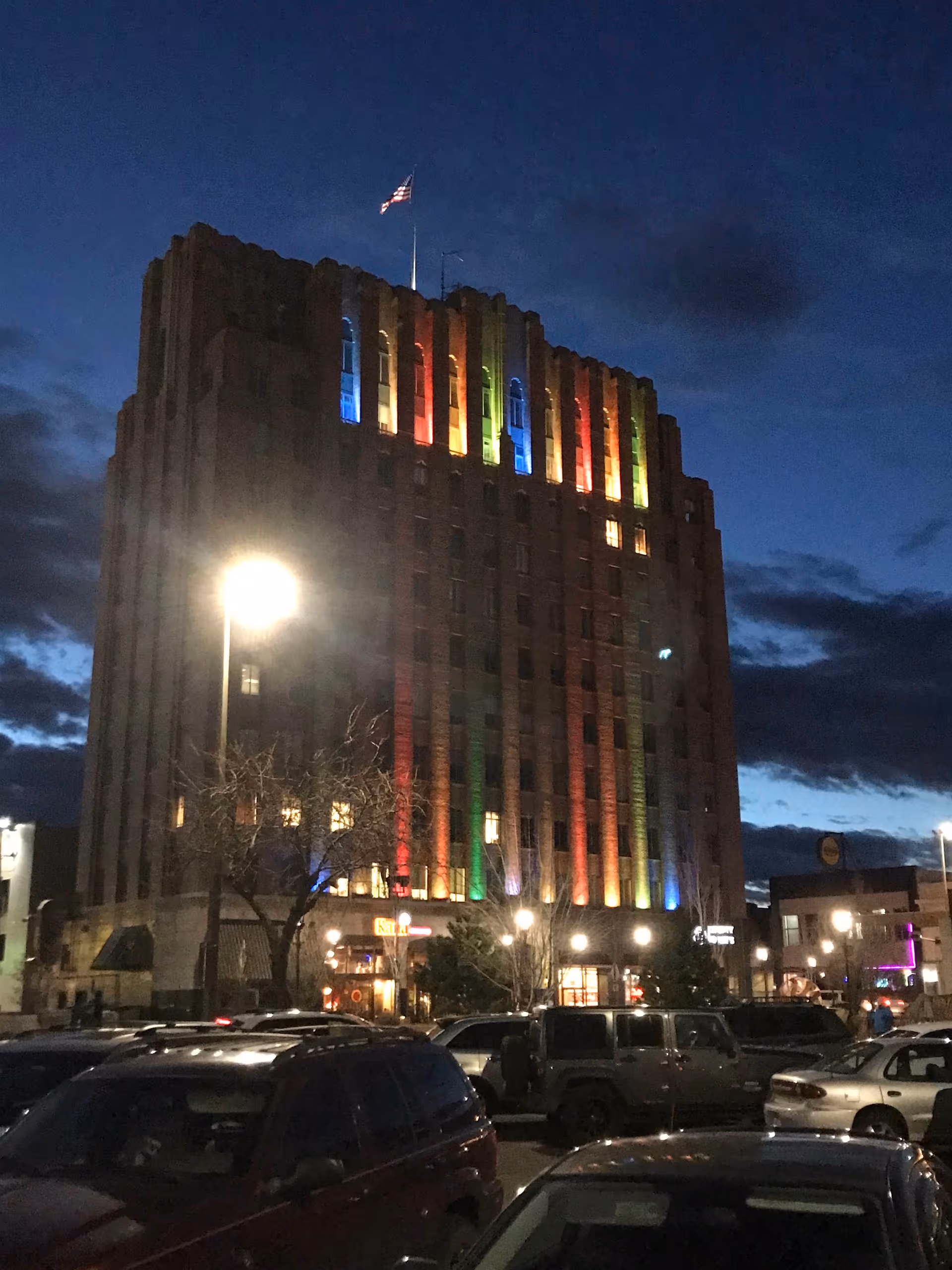 Nighttime city street view of a tall rainbow-lit building with parked cars in foreground.