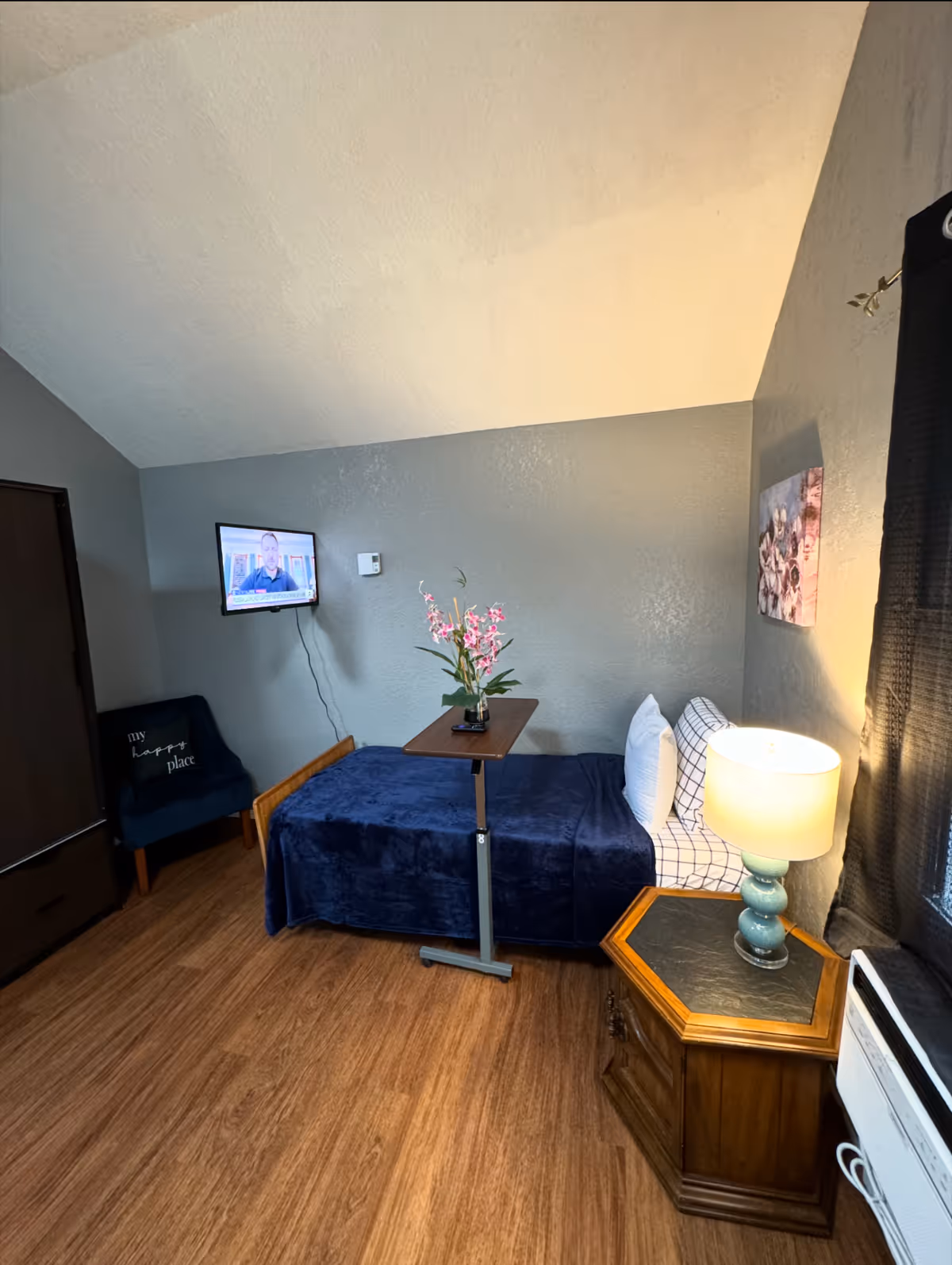 A compact bedroom at Empathy Haven AFH LLC with a bed, TV, nightstand, lamp, and flowers.